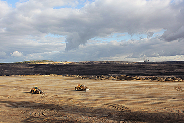 Weitläufige Landschaft eines aktiven oder ehemaligen Braunkohletagebaus in der Lausitz. Im Vordergrund zwei Baumaschinen auf sandigem, weit aufgerissenem Gelände mit deutlichen Fahrspuren. In der Ferne sind dunkle Erdschichten und ein großer Schaufelradbagger zu sehen. Der Himmel ist von dramatischen Wolken durchzogen, einzelne Sonnenstrahlen brechen durch.