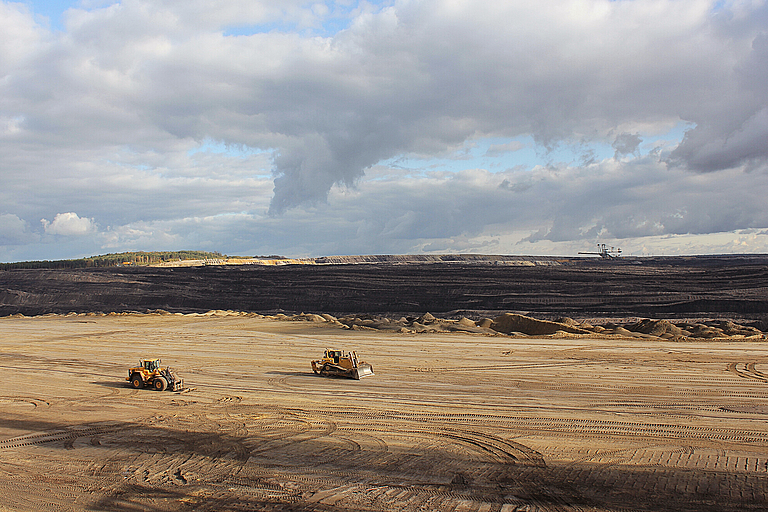 Weitläufige Landschaft eines aktiven oder ehemaligen Braunkohletagebaus in der Lausitz. Im Vordergrund zwei Baumaschinen auf sandigem, weit aufgerissenem Gelände mit deutlichen Fahrspuren. In der Ferne sind dunkle Erdschichten und ein großer Schaufelradbagger zu sehen. Der Himmel ist von dramatischen Wolken durchzogen, einzelne Sonnenstrahlen brechen durch.